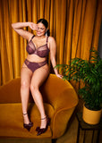 Woman in a burgundy lingerie set sitting on an orange chair with a plant in the background.